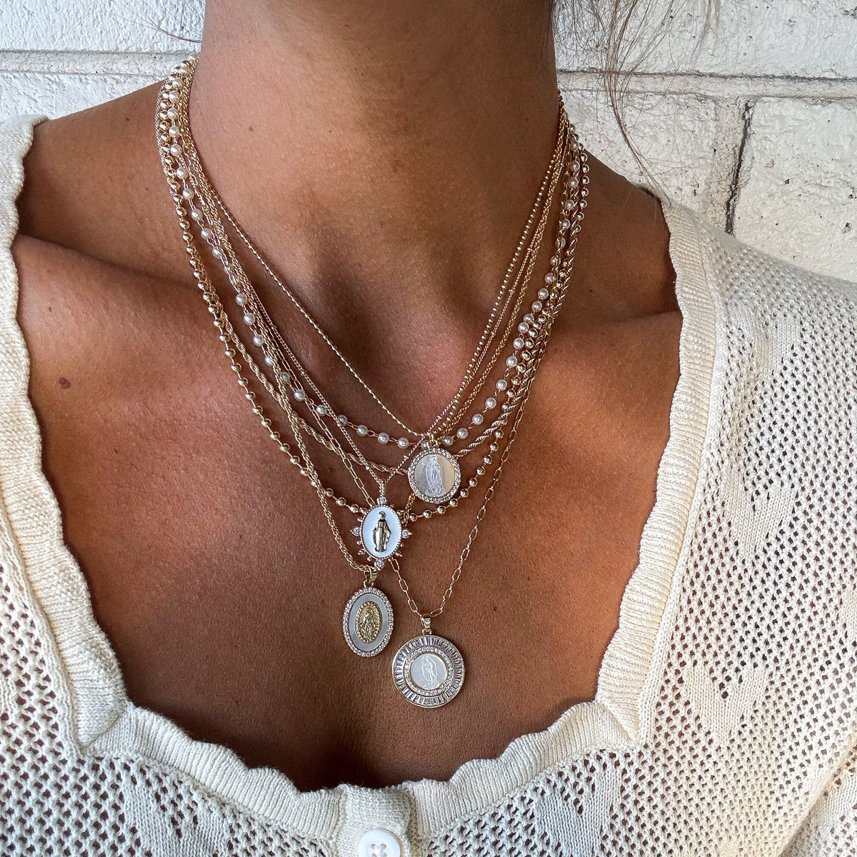 Layered gold-tone necklaces with Marian pendants, worn with a white knit top